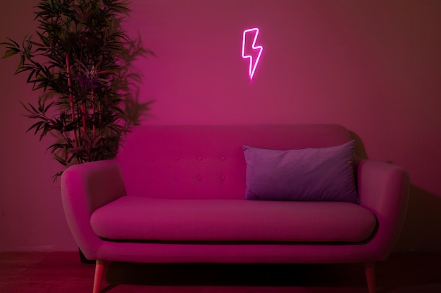 Розовый диван с фиолетовой подушкой стоит под неоновой вывеской в ​​виде молнии рядом с растением в горшке.