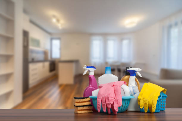 Чистящие средства, включая распылители, перчатки и губки, на столе в светлой современной кухне.